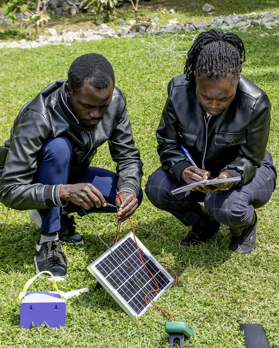 Two students outdoors wiring a solar panel to the EMA Power Box, one taking notes